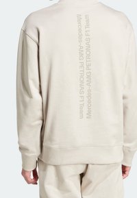 Beige mikina a kalhoty s vertikálním nápisem "Mercedes-AMG PETRONAS F1 Team" potisknutým uprostřed zad mikiny.