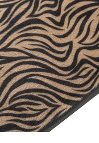 Tappeto color nocciola con motivi zebrati neri ondulati, texture morbida e bordi dritti. La superficie è pelosa, migliorando il contrasto visivo del design.