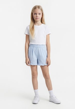 Mlada deklica z dolgimi blond lasmi, oblečena v belo majico, svetlo modre črtaste hlače z volani na robu, bele nogavice in bele superge, stoji.