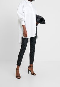 Persona que lleva una camisa blanca larga con botones, pantalones negros con lunares, sandalias de tacón negro, sosteniendo una chaqueta de cuero negra doblada.