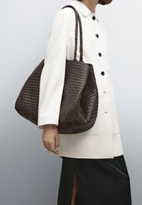 Woven dark brown tote bag with two braided handles. Slouchy shape with a textured surface. Model wearing a white jacket and black skirt.