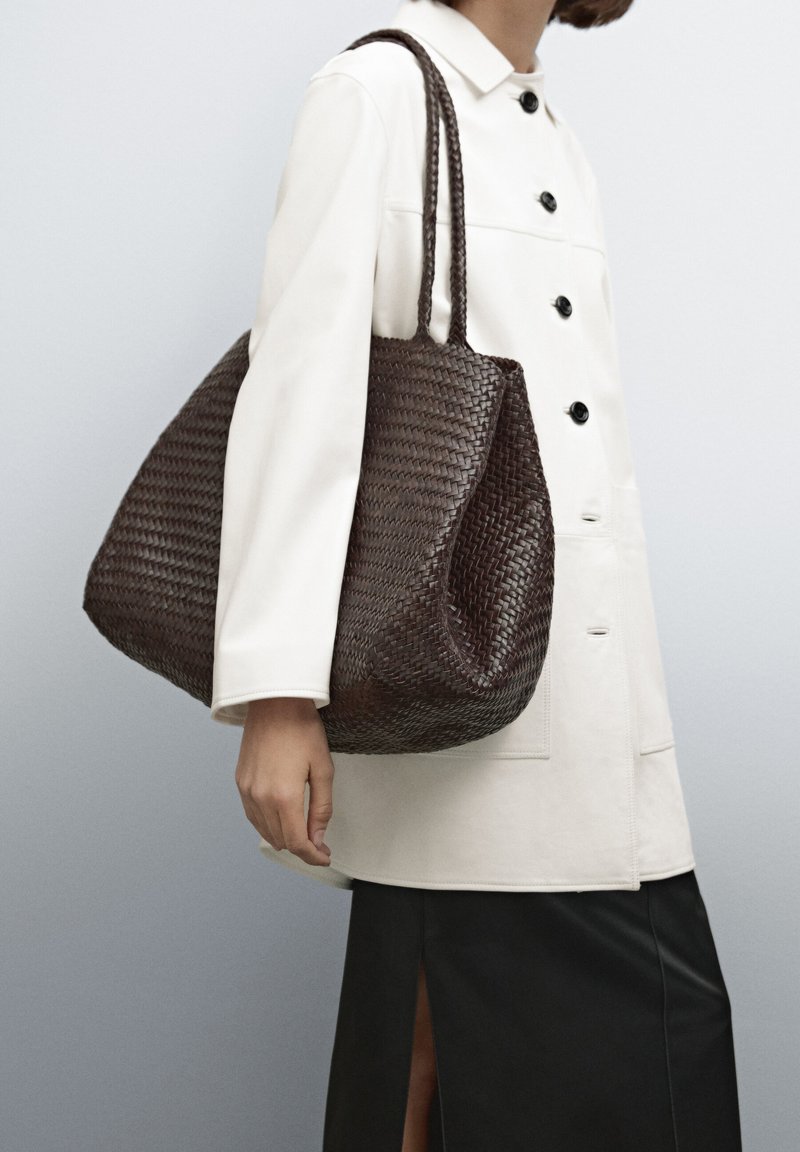 Woven dark brown tote bag with two braided handles. Slouchy shape with a textured surface. Model wearing a white jacket and black skirt.