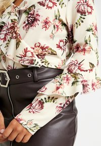 Blusa floreale con sfondo crema, caratterizzata da maniche volant, in tonalità di bordeaux e verde. Abbinata a pantaloni di pelle marrone scuro e una cintura argentata.