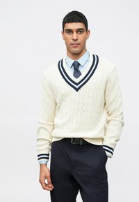 Σουέτερ σε κρέμα από πλεκτό με V-λαιμόκοψη, διακοσμημένο με ναυτικές και λευκές ρίγες, συνδυασμένο με ανοιχτό μπλέ πουκάμισο και σκούρους παντελόνες.