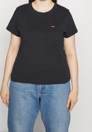 Kobieta w prostym, czarnym t-shircie Levi's z krótkim rękawem i jasnoniebieskich dżinsach, stojąca na jednolitym tle.