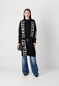 Črn dolg kardigan z belim napisom "DKNY", v kombinaciji s črnim topom in širokimi modrimi džinsi. Dodatki vključujejo majhno črno ročno torbico.