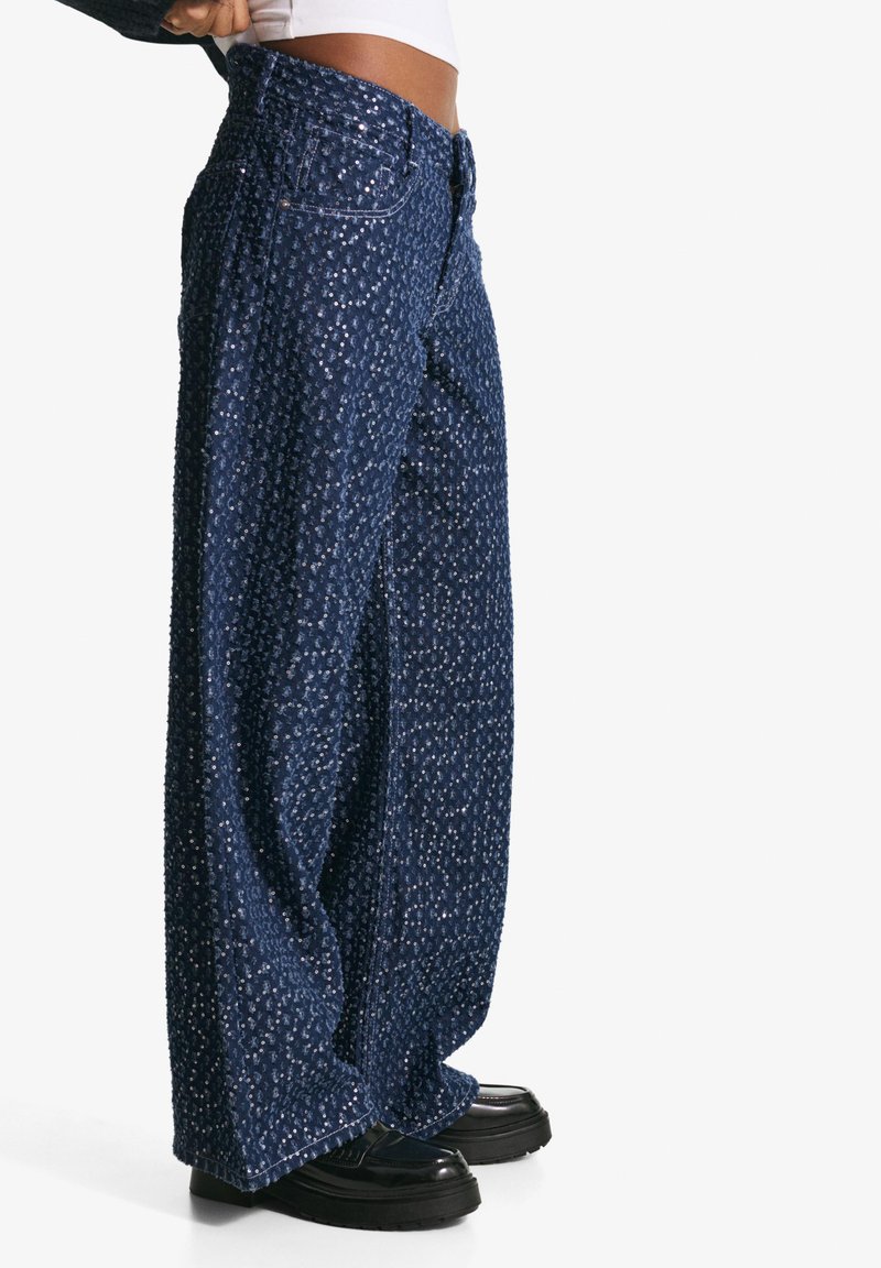 Široke traperice u tamnoplavoj boji s teksturiranim uzorkom, s džepovima i zatvaranjem na gumb. Nositi s glomaznim crnim platformama.