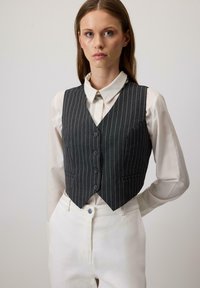 Vrouw in een donkergrijs gestreept vest over een wit geknoopte shirt en witte broek, staande tegen een lichte effen achtergrond.