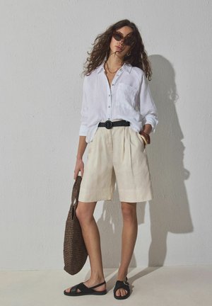 Žena s kudrnatými vlasy, oblečená v nadměrné bílé košili na zapínání, béžových šortkách ke kolenům, černých sandálech, slunečních brýlích a držící pletenou hnědou tašku.