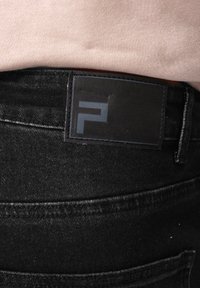 Asmuo dėvi juodus džinsus su juodu stačiakampiu pleistru juosmens srityje, kuriame yra minimalistinis pilkas logotipas, derinamus su šviesiai rožine palaidine.