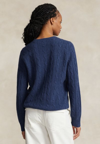 Naine palmikutega juustega, kes kannab sinist palmikutega kampsunit ja valgeid pükse, pööratud seljaga neutraalse tausta ees.