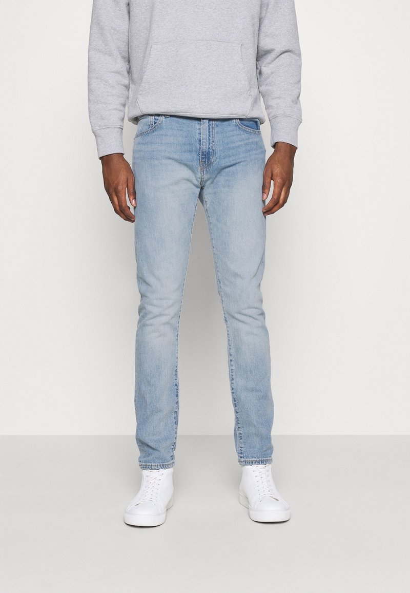 Homme portant un jean slim bleu clair, un sweat à capuche gris et des baskets blanches, debout devant un fond blanc uni.