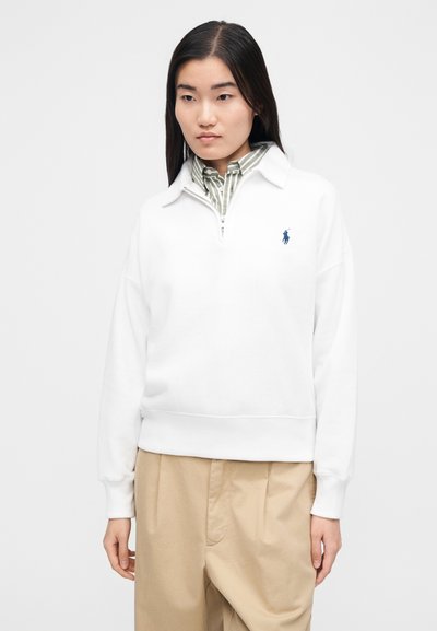 Sudadera blanca con cremallera y cuello con solapa, con un pequeño logo azul en el pecho izquierdo y puños acanalados. Llevada sobre una camiseta de rayas verdes.