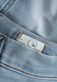 Lys blå denim med synlige sting. Har en hvit midjebåndsetikett og en klar knapp. Teksturen ser myk og litt slitt ut.