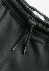 Dunkelgrüne Sweatpants aus strukturiertem Material, mit elastischem Bund und einem geknoteten Kordelzug, der schwarze Metallspitzen aufweist. Etikett sichtbar.