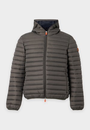 Homme portant une veste matelassée à capuche vert olive sur une chemise blanche, avec un pantalon large noir et des baskets blanches, debout devant un fond uni.