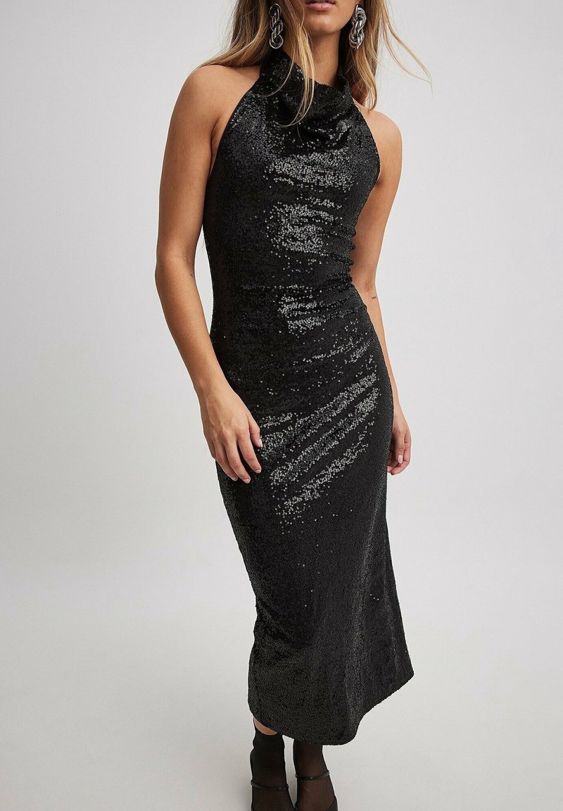 Femme portant une robe midi noire sans manches à col bénitier ornée de sequins, avec des bottines noires et de grandes boucles d'oreilles argentées, debout devant un fond uni.
