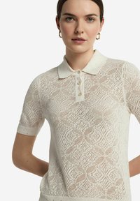 Pikétröja i off-white, med ett texturerat spetsmönster, korta ärmar och en knappkrage med små dekorativa inslag.