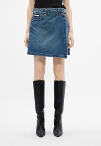 Denim-Minirock in einem mittleren Blauton, mit einer Vordertasche, asymmetrischem Saum und genähten Details. Kombiniert mit knielangen schwarzen Stiefeln.