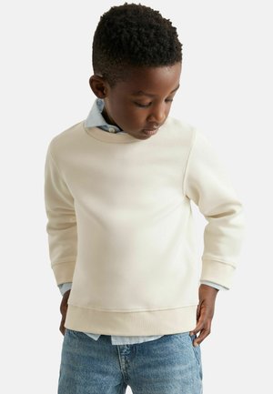 Balta kokvilnas sporta jaka ar tekstūru, apaļo kakla izgriezumu un ribotām piedurknēm, uzvilkta virs gaiši zila apkakles krekla, kombinēta ar džinsu biksēm.