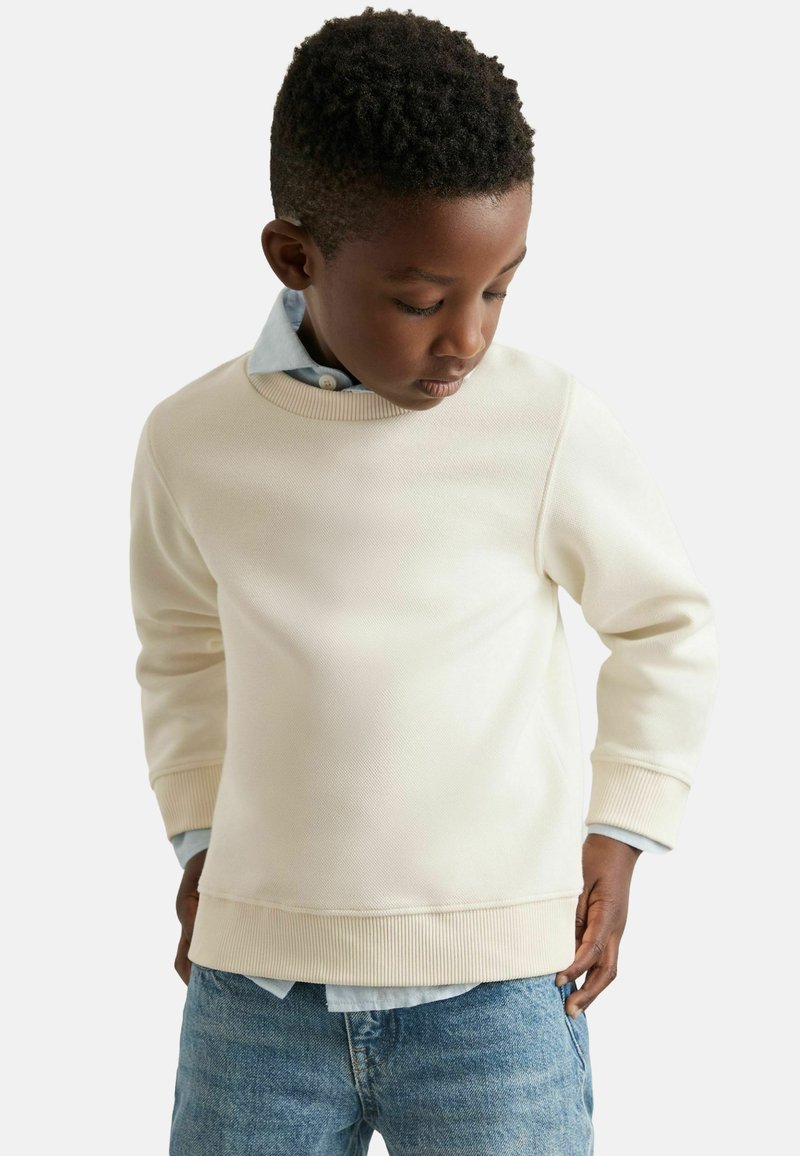 Valge puuvillane dressipluus tekstureeritud viimistlusega, ümar kaelus ja soonikvarrukad, kantud helesinise kraega särgi peale, koos teksadega.