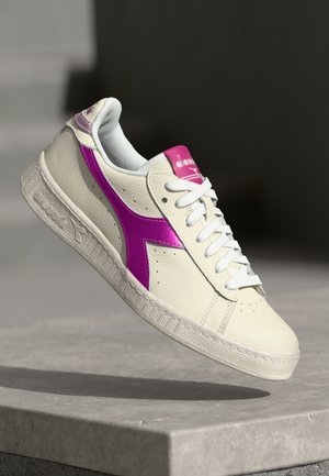Baskets en cuir blanc avec un logo violet sur le côté, lacées de blanc et portant une étiquette rose sur la languette, suspendues au-dessus d'une surface en pierre grise.