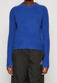 Blauer gestrickter Pullover mit rundem Halsausschnitt, gerippter Textur und langen Ärmeln; leicht verkürzter Saum. Kombiniert mit schwarzen Hosen.