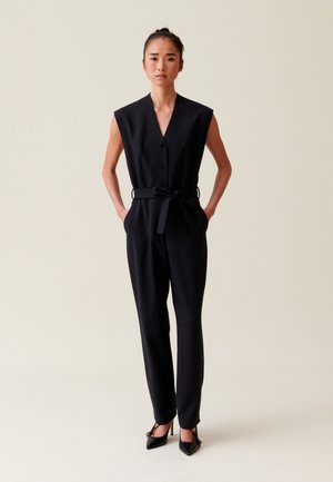 Combinaison noire avec un décolleté en V, sans manches, fermeture à boutons sur le devant et ceinture nouée à la taille. Tissu lisse et structuré ; le modèle porte des talons hauts.