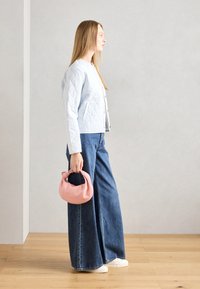 Veste matelassée bleu clair avec un devant boutonné, associée à un jean en denim bleu à jambe large. Tenant un sac à main en tissu rose. Fond en parquet.