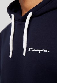 Sweat à capuche bleu marine avec cordons blancs et logo blanc "Champion" imprimé sur la poitrine, porté par une personne.