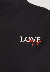 Tissu noir avec texte blanc "LOVE" et "Hugo" rouge brodé en cursive superposé aux lettres sur la partie supérieure de la poitrine d'un vêtement.