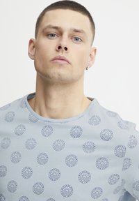 Lyseblå T-skjorte laget av bomull, med et mønster av sirkulære logoer i mørkeblått over hele, rund halsdesign og korte ermer.