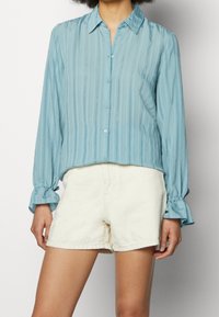 Hellblaues, gestreiftes Hemd mit langen Ärmeln und gerafften Manschetten, getragen mit cremefarbenen Shorts, präsentiert an einem Modell vor einem schlichten Hintergrund.