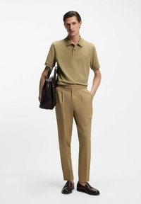 Koszulka polo w kolorze khaki z kołnierzykiem, połączona z dopasowanymi beżowymi spodniami i brązowymi mokasynami. Modelka trzyma ciemnobrązową torbę typu tote.