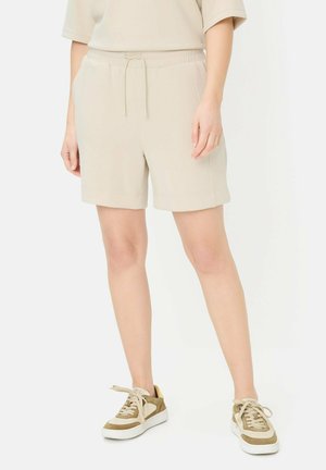 Personne portant un short beige mi-long à taille ajustable avec un cordon et des baskets assorties à semelles blanches, debout devant un fond blanc.