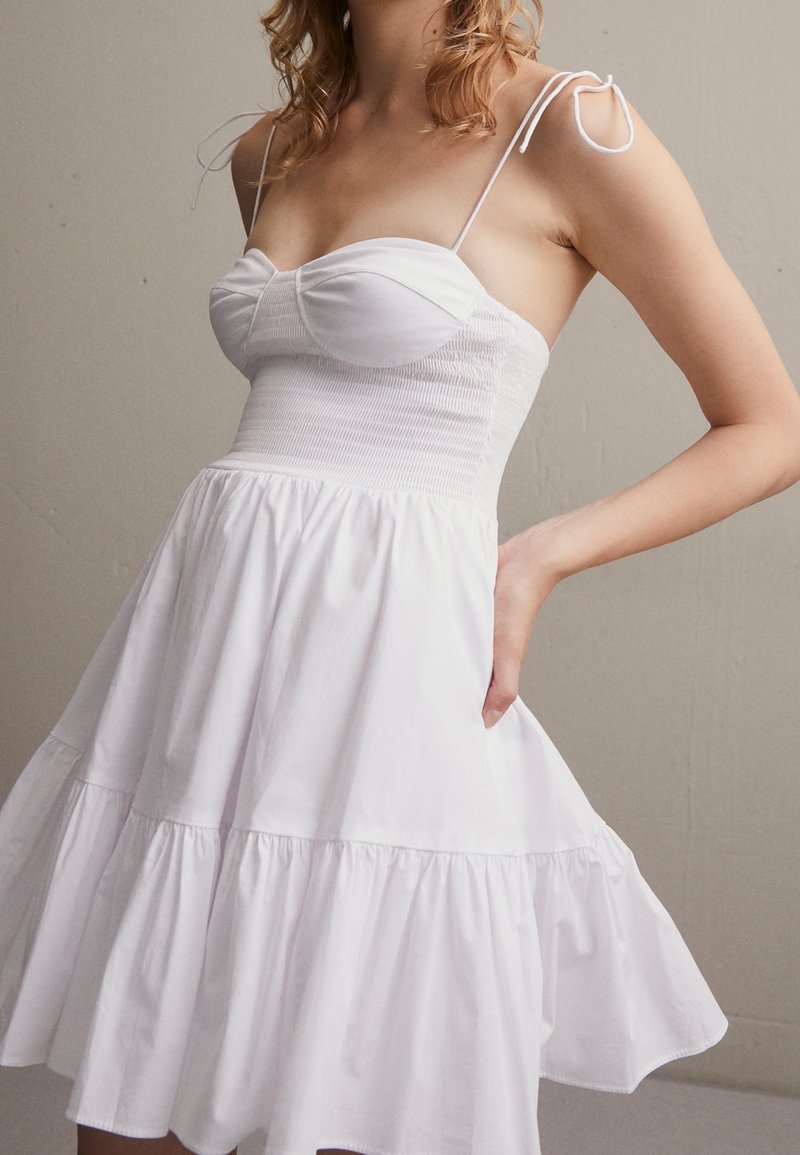 Bílá bavlněná šaty s přiléhavým živůtkem, nabíranou texturou a nastavitelnými ramínky. Rozšířená sukně s patkou, poskytující volný střih.