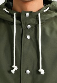 Olivově zelená bunda s kapucí, se stříbrnými patentkami a bílými šňůrkami. Hladká textura s minimalistickým designem.