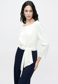 Hvit, langermet topp med rund hals og knytt detalj i livet, kombinert med marineblå bukser, som viser en glatt tekstur på stoffet.