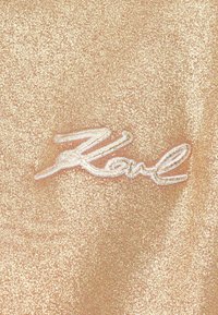 Material auriu sclipitor cu o suprafață texturată, având o semnătură brodată albă "Karl" centrală în design.