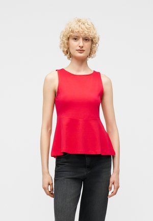 Kvinde med kort krøllet blondt hår iført en ærmeløs rød peplum-top og sorte jeans, stående foran en ensfarvet lys baggrund.