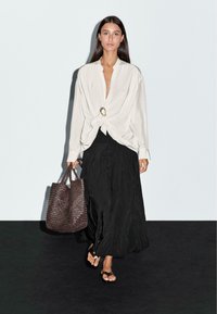 Donna che cammina indossando una blusa bianca a maniche lunghe con una chiusura dorata, una gonna maxi nera, sandali neri e portando una grande borsa a tracolla intrecciata marrone.