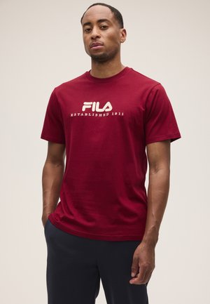 Uomo che indossa una T-shirt rossa FILA con il testo "Established 1911", in piedi con una mano in tasca contro uno sfondo chiaro e uniforme.