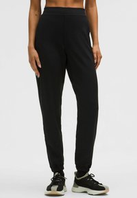 Juodi sportiniai kelnės su lygiu paviršiumi, turinčios siaurėjantį siluetą, elastinį juosmenį ir apvalkalus ant kulkšnių, derinamos su juodais ir pilkais sportbačiais.