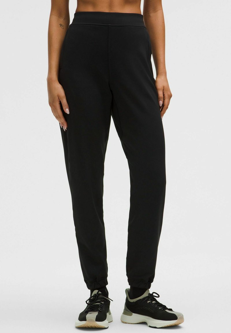 Juodi sportiniai kelnės su lygiu paviršiumi, turinčios siaurėjantį siluetą, elastinį juosmenį ir apvalkalus ant kulkšnių, derinamos su juodais ir pilkais sportbačiais.