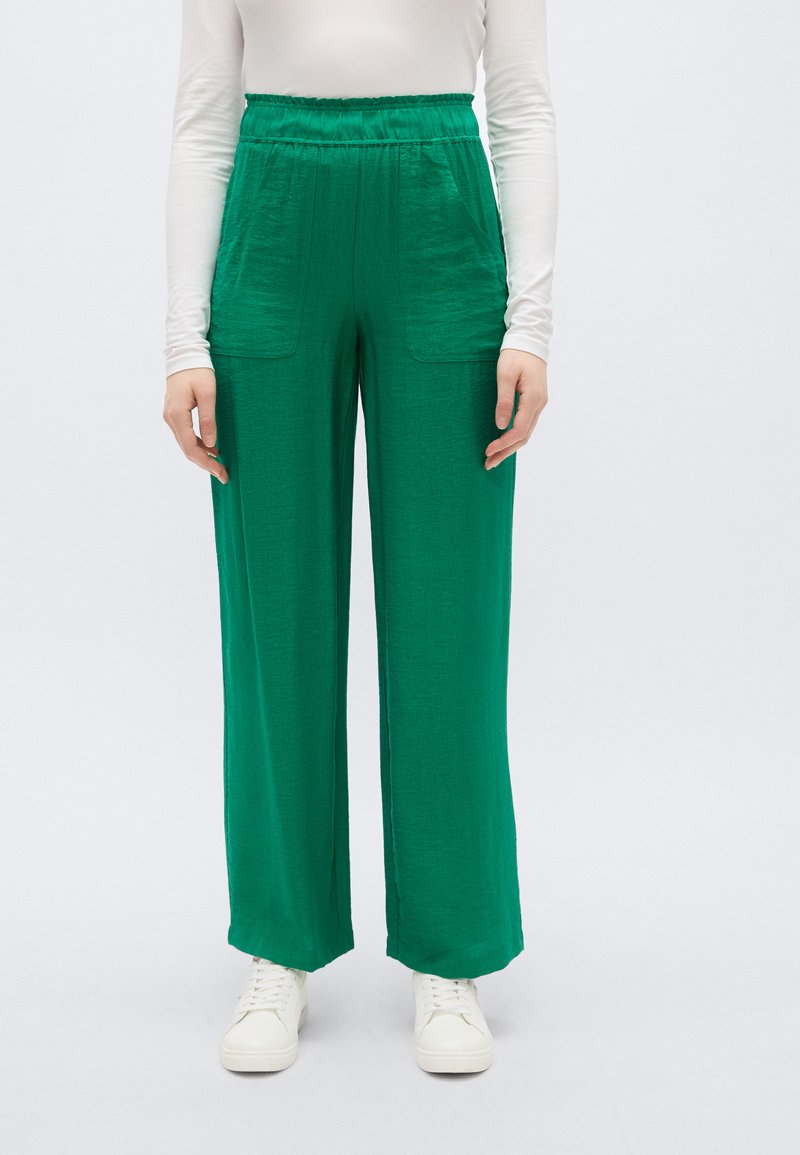 Πράσινο παντελόνι με φαρδιά γραμμή και υφή, με δύο τσέπες μπροστά και ελαστική μέση. Συνδυάζεται με λευκά αθλητικά παπούτσια.