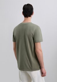 Man met kort, donker haar, gekleed in een olijfgroen T-shirt en witte broek, staand met zijn gezicht naar weg van een effen lichtgrijze achtergrond.