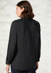 Maglietta nera a maniche lunghe con una texture liscia, polsini abbottonati e una vestibilità rilassata. Presenta un dettaglio di cucitura sulla spalla posteriore e una morbida caduta.