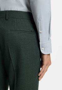 Donkergroene, gestructureerde broek gecombineerd met een lichtblauw, glad jurkhemd met knoopmanchetten en een eenvoudige zakdetail.