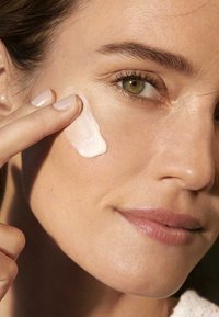 Crème appliquée sur la joue, texture lisse, couleur rose clair. Gros plan sur le teint de la peau, l'œil, et placement délicat de la main.