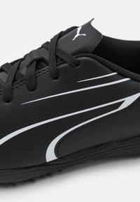 Scarpa sportiva nera con tomaia sintetica, strisce bianche a contrasto e suola in gomma testurizzata con piccoli chiodi per la trazione.